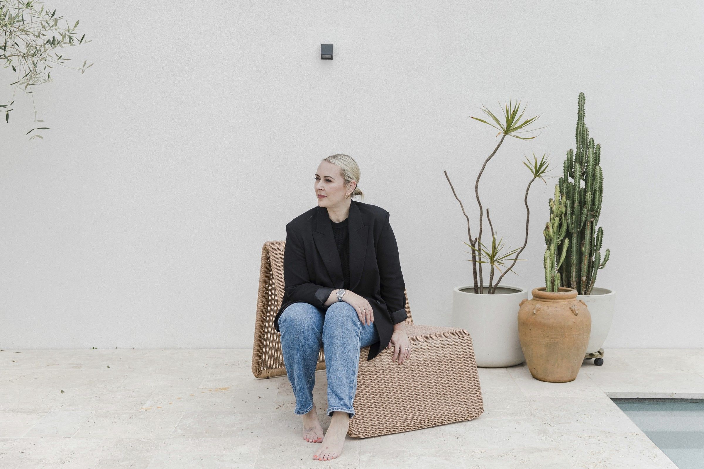 Jess sitting on a woven chair against a white wall, beside potted plants